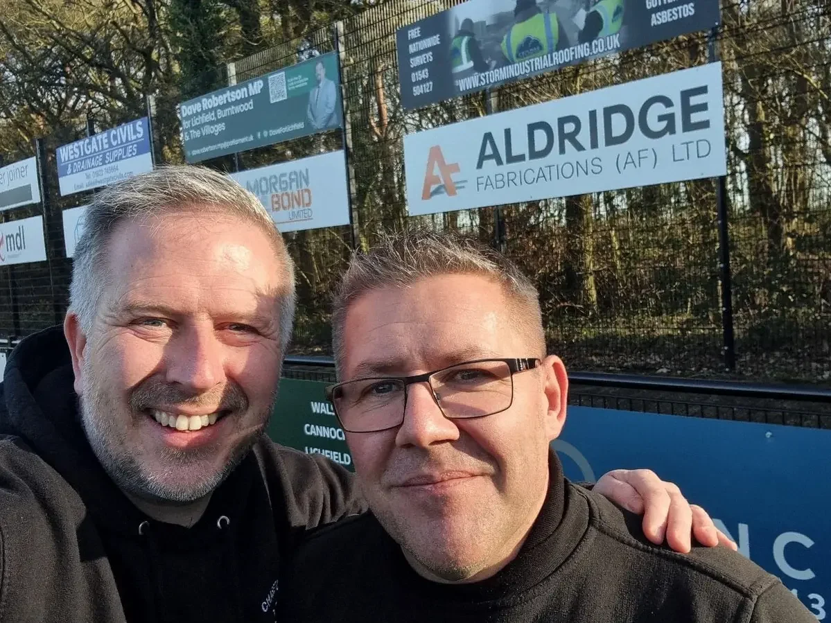 Darren Whitehouse from Aldridge Fabrications standing pitchside at Chasetown FC with the AF sponsor board in the foreground and supporter flags in the main stand